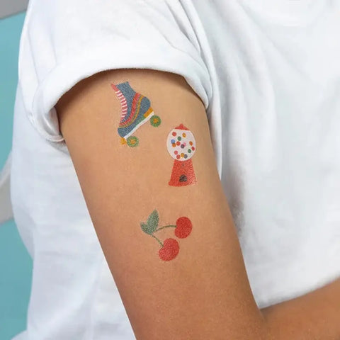 Eine Person trägt ein weißes T-Shirt und zeigt drei witzige Tattoos - einen Rollschuh, einen Kaugummiautomaten und Kirschen - perfekte Kindertattoos aus den Abwaschbaren Tattoos - Top Banana (2 Bögen) von Rex London.