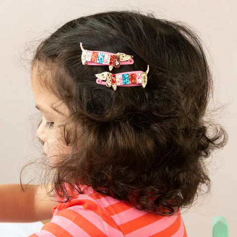 Ein Kind mit lockigem, dunklem Haar trägt eine rosa gestreifte Bluse und hat zwei "Dackel" Glitzer-Haarspangen von Rex London seitlich im Haar - eine tolle Geschenkidee für Kinder. Das Profil hebt sich vor hellem Hintergrund ab.