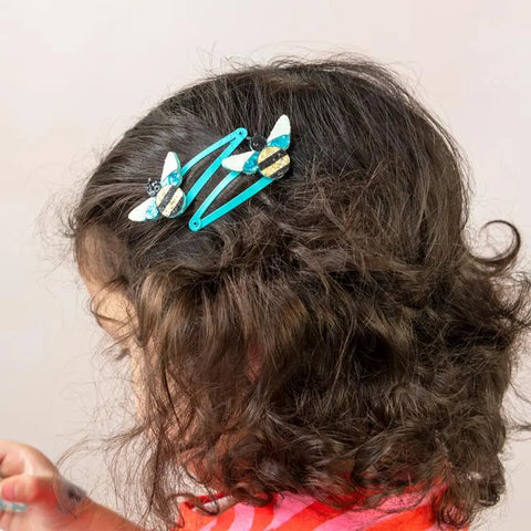 Ein Kind mit lockigem braunem Haar trägt links zwei "Hummel" Glitzer-Haarspangen (2er Set) von Rex London, im Profil gesehen in einem rot gemusterten Oberteil - perfekt für süße Geschenkideen für die Kleinen.