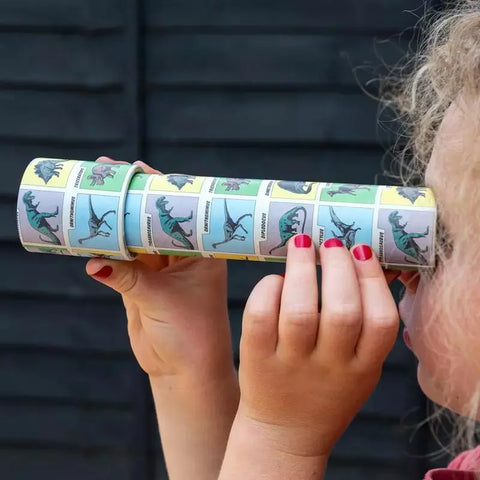 Ein Kind mit lockigem Haar und rotem Nagellack blickt durch das Kaleidoskop Prehistoric Land von Rex London mit Dinosaurier-Illustrationen - ein ideales Geschenk für Kinder -, das es sich mit beiden Händen vor die Augen hält.