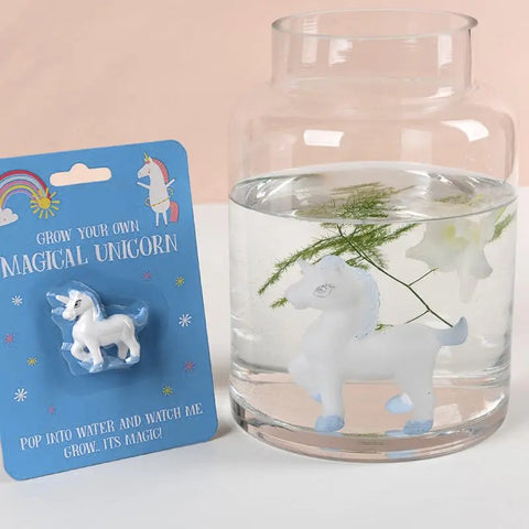 Ein kleines Magisches Einhorn von Rex London sitzt neben einem Glas mit Wasser, das seine größere, erweiterte Form und einen Zweig mit Grünzeug enthält.