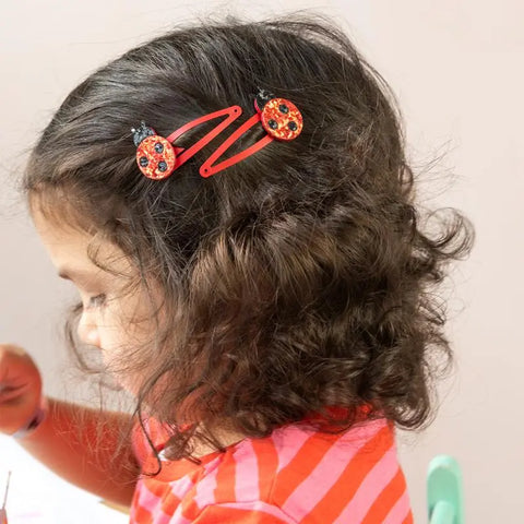 Ein junges Mädchen mit lockigem braunem Haar, im Profil gesehen und auf eine unsichtbare Tätigkeit konzentriert, trägt ein rosa-rot gestreiftes Hemd und zwei Rex London "Marienkäfer" Glitzer-Haarspangen (2er Set) - eine charmante Geschenkidee.