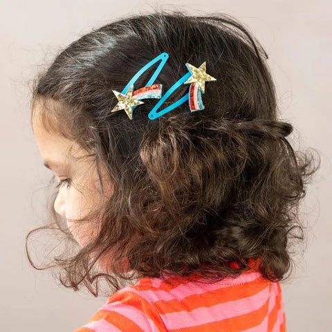 Ein junges Mädchen mit gewellten braunen Haaren trägt ein rot-rosa gestreiftes Hemd und zwei "Sternschnuppe" Glitzer-Haarspangen (2er Set) von Rex London - verziert mit goldenen Glitzersternen, Bändern und perfekt als Sternschnuppen Geschenkideen - während es nach links schaut.
