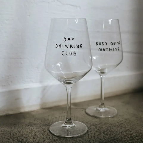 Zwei "Day Drinking Club" Weingläser von Johanna Schwarzer für Selekkt: Eines mit "DAY DRINKING CLUB"-Print, das andere mit "BUSY DOING NOTHING". Beide Allrounder sind handbeschriftet und stehen auf Beton vor einer weißen Wand.