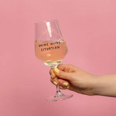 Eine Hand mit gelb lackierten Nägeln hält das mit Weißwein gefüllte Weinglas "Wine Wine Situation" von Johanna Schwarzer (Selekkt). Das Glas trägt seinen Namen auf einem einfarbig rosa Hintergrund.