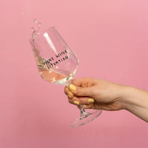 Eine Hand mit gelben Nägeln hält ein gekipptes "Wine Wine Situation" Weinglas von Johanna Schwarzer für Selekkt und verschüttet Weißwein. Das verspielte Glas mit dem aufgedruckten Slogan hebt sich von einem rosafarbenen Hintergrund ab - ein aufmerksamkeitsstarker Moment.