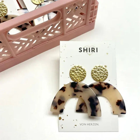 Ein Paar Shiri Ohrringe "Berlin" mit goldenen texturierten runden Nieten und großen gewölbten Schildpatt-Anhängern, abgebildet auf einer weißen Karte neben einem rosa perforierten Korb - ideal als Statement-Schmuck.