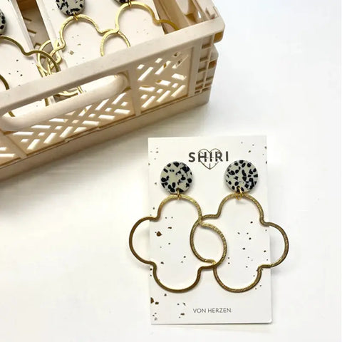 Ein Paar Ohrringe "Casablanca" von Shiri Ohrringe mit runden weiß-schwarz gesprenkelten Ohrsteckern und großen abstrakten Golddrahtformen, ausgestellt auf einer Shiri-Karte neben einer beigen Kiste mit mehr Schmuck aus Hannover.