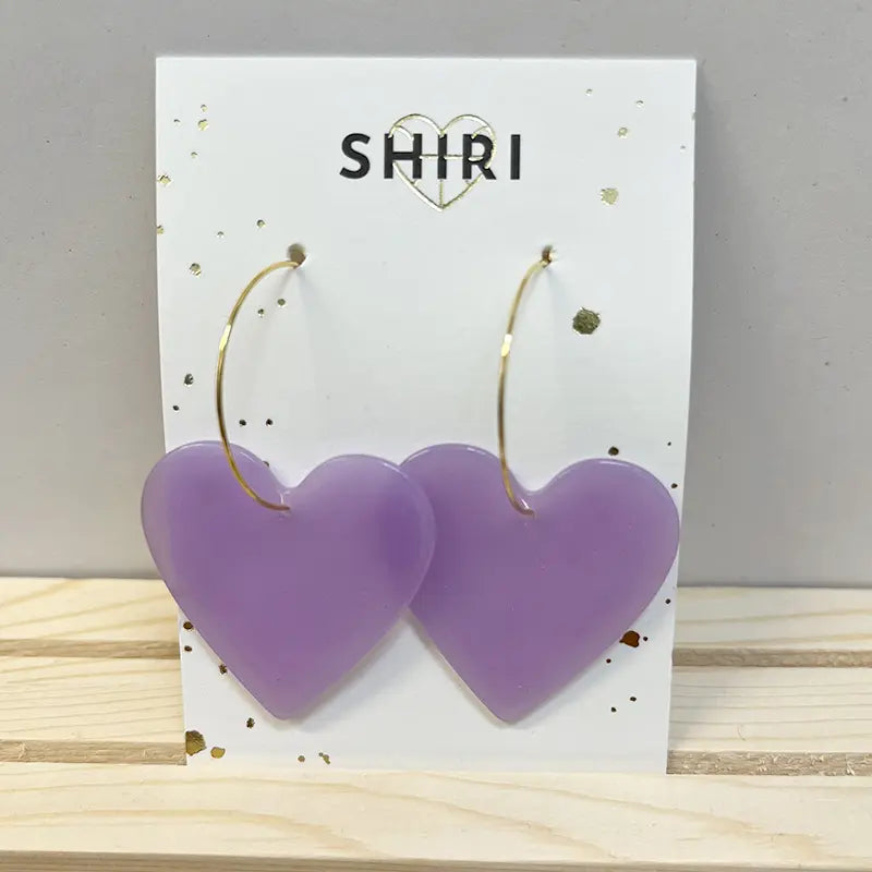 Ein Paar große, lilafarbene Ohrringe in Herzform "Havanna" von Shiri mit dünnen Goldreifen wird auf einer weißen SHIRI-Karte vor hellem Holz und einer grauen Wand präsentiert und verleiht jedem Look einen lebendigen Charme.