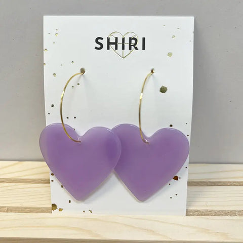 Ein Paar große, lilafarbene Ohrringe in Herzform "Havanna" von Shiri mit dünnen Goldreifen wird auf einer weißen SHIRI-Karte vor hellem Holz und einer grauen Wand präsentiert und verleiht jedem Look einen lebendigen Charme.