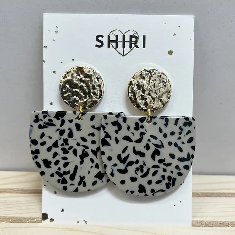 Ein Paar Shiri Ohrringe "Kairo" mit runden, strukturierten Goldnieten und großen, schwarz-weiß gesprenkelten Halbmond-Anhängern - Statement-Schmuck von Shiri, präsentiert auf weißer Karte mit SHIRI-Logo und Herz.