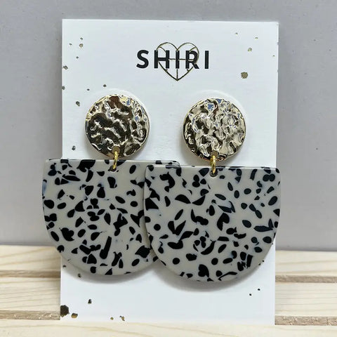 Ein Paar Shiri Ohrringe "Kairo" mit runden, strukturierten Goldnieten und großen, schwarz-weiß gesprenkelten Halbmond-Anhängern - Statement-Schmuck von Shiri, präsentiert auf weißer Karte mit SHIRI-Logo und Herz.