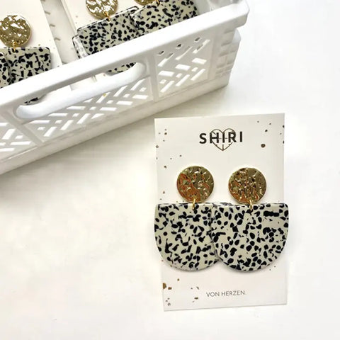 Ein Paar "Kairo" von Shiri Ohrringe mit goldenen, runden Ohrsteckern und schwarz-weiß gesprenkelten, halbkreisförmigen Anhängern wird auf einer Markenkarte neben einem weißen Plastikkorb mit weiteren Schmuckstücken präsentiert.