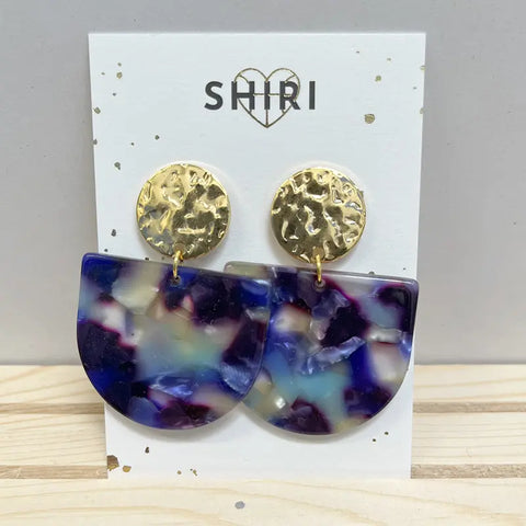 Ein Paar Shiri Ohrringe "Kopenhagen" mit gehämmerten goldfarbenen Nieten und großen marmorierten violetten, blauen und weißen Tropfen - perfekt als Statement-Schmuck in Hannover - ausgestellt auf einer Karte mit der Aufschrift SHIRI.