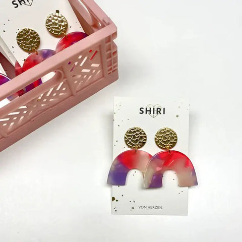 Ein Paar farbenfrohe, halbrunde Ohrringe "Las Vegas" von Shiri mit goldenen, strukturierten Ohrsteckern ruht auf einer Ausstellungskarte mit der Aufschrift Shiri, neben einer rosafarbenen Kiste mit weiteren Statement-Schmuck-Ohrringen desselben Designs.