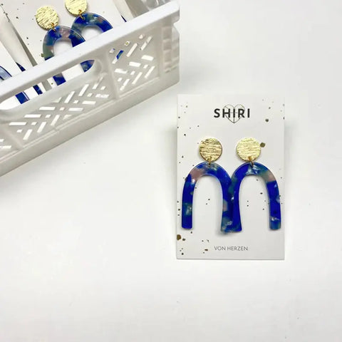 Ein Paar blau-goldene Shiri Ohrringe "Los Angeles" in Bogenform auf einer weißen SHIRI-Markenkarte, daneben ein dekorativer Korb mit weiteren Acetat-Schmuckstücken - perfekt für Statement-Schmuck Hannover.