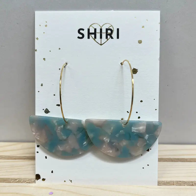 Ein Paar Ohrringe "Nizza" von Shiri mit großen goldenen Reifen und blau-weiß marmorierten Acetat-Anhängern, die auf einer Markenkarte mit einem goldenen Herz über dem Namen präsentiert werden.