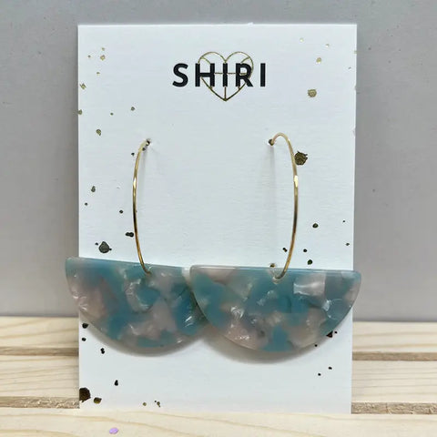Ein Paar Ohrringe "Nizza" von Shiri mit großen goldenen Reifen und blau-weiß marmorierten Acetat-Anhängern, die auf einer Markenkarte mit einem goldenen Herz über dem Namen präsentiert werden.