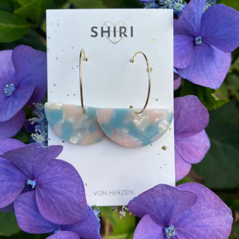 Ein Paar Ohrringe "Nizza" von Shiri in pastellblauen und rosafarbenen Acetat-Halbkreisen, ausgestellt auf einer Shiri-Karte inmitten leuchtend lila Hortensienblüten.