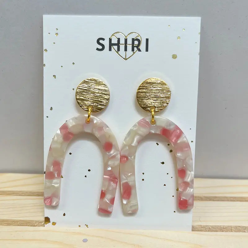 Die Ohrringe "Orlando" von Shiri bestehen aus strukturierten, runden goldenen Ohrsteckern und U-förmigen Acetat-Ohrringen in transluzentem Rosa und Weiß, die auf einer Shiri-Markenkarte präsentiert werden. Schmuck aus Hannover.