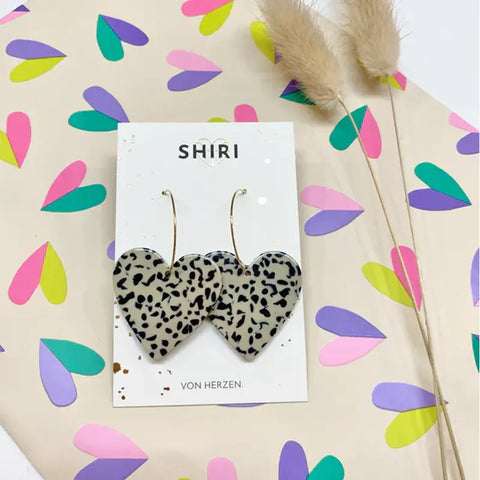 Ein Paar herzförmige, schwarz-weiße Ohrringe mit Leopardenmuster "Rio" von Shiri auf einer Markenkarte, auf blütengemustertem Papier mit beigen Trockengräsern. Leichte, handgemachte Ohrringe aus Hannover.