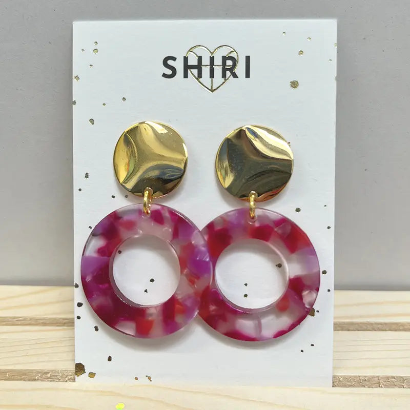 Ein Paar Shiri Ohrringe "Team Bold" mit glänzenden runden Goldnieten und großen marmorierten rosa und lila Harzreifen, präsentiert auf einem weißen SHIRI card-trendiger Statement-Schmuck aus Hannover.