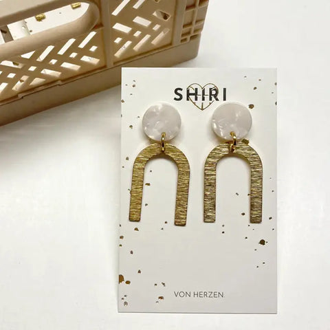 Die Ohrringe "Wien" von Shiri zeichnen sich durch runde, marmorierte weiße Spitzen und große, strukturierte Goldbögen aus, die auf einer weißen Karte mit goldenen Sprenkeln und dem eleganten Markennamen Schmuck aus Hannover präsentiert werden.