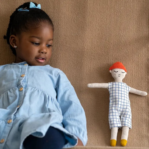 Ein junges Mädchen in einem hellblauen Kleid sitzt auf einer braunen Fläche und schaut auf die Sophie Home Stoffpuppe "Buddy Grid" aus Baumwolle neben sich. Die Puppe hat orangefarbene Haare, ein kariertes Outfit und gelbe Schuhe.