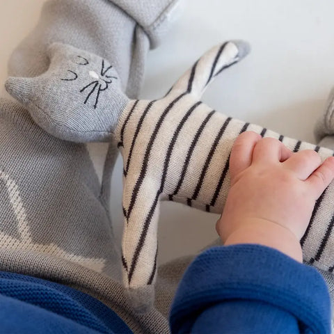 Ein Baby in Blau und Grau hält sitzend eine weiche, gestreifte, grau-weiße Stoffpuppe "Katze" Baumwolle von Sophie Home.