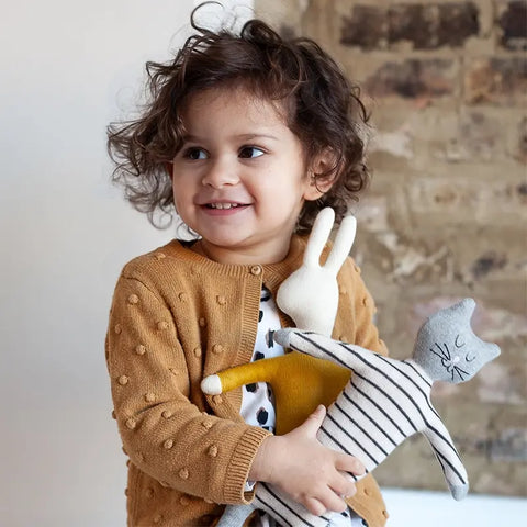 Ein kleines Kind mit lockigem Haar, brauner Strickjacke und gepunktetem Kleid lächelt, während es zwei Sophie Home Stoffpuppe "Katze" Baumwolle-Spielzeuge - ein weißes Kaninchen und eine grau gestreifte Katze - vor einem Hintergrund aus Backsteinmauern hält.