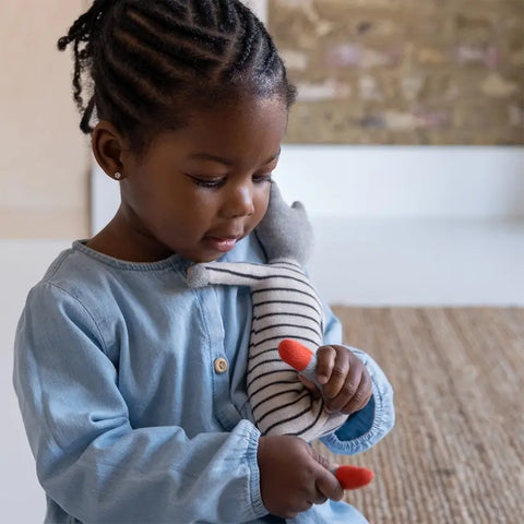 Ein kleines Kind mit geflochtenen Haaren und einem blauen Kleid hält eine Sophie Home Stoffpuppe "Katze" Baumwolle und berührt sanft ihre roten Füße, während es auf einem gewebten Teppich in einem sanft beleuchteten Raum sitzt.