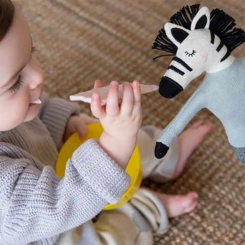 Ein kleines Kind in einem grauen Pullover sitzt auf einem gewebten Teppich, hält eine gelbe Schale in der Hand und tut so, als würde es eine Sophie Home Stoffpuppe "Zebra" Baumwolle mit einem Spielzeuglöffel füttern.