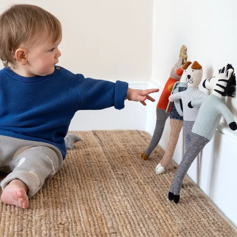 Ein Kleinkind in einem blauen Pullover und einer grauen Hose sitzt auf einem Teppich und greift nach der Sophie Home Stoffpuppe "Zebra" Baumwolle, während drei andere Plüschpuppen an der Wand aufgereiht sind.