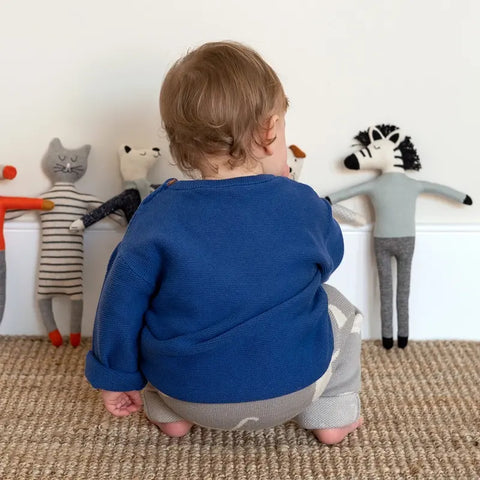 Ein kleines Kind in einem blauen Baumwollstrick-Pullover und einer grauen Hose hockt auf einem gewebten Teppich, von der Kamera abgewandt, und spielt mit dem Plüschtier Sophie Home Stoffpuppe "Zebra" Baumwolle, das vor einer weißen Wand steht.
