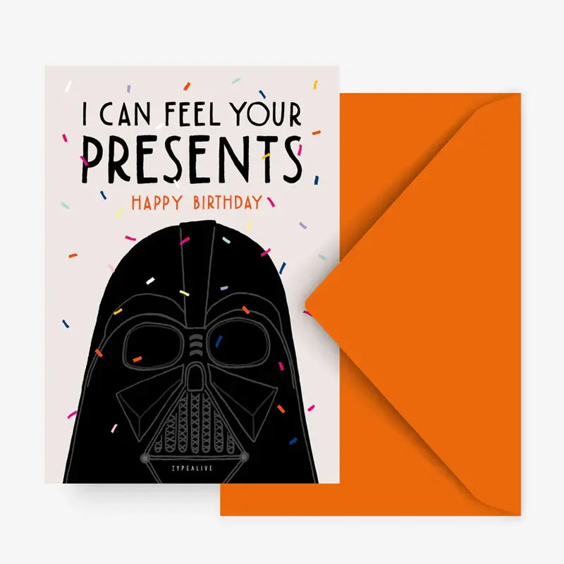 Die Postkarte "V*der's Birthday Wishes" von Typealive zeigt eine maskierte dunkle Figur mit Konfetti und dem Text "I CAN FEEL YOUR PRESENTS. HAPPY BIRTHDAY." Ein orangefarbener Umschlag vervollständigt dieses einzigartige Design.
